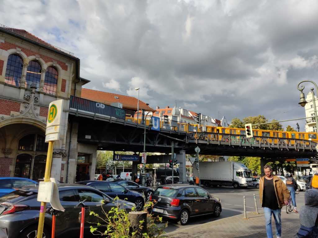 Cars are stuck near Schlesisches Tor. Above the yellow trainsystem called RING-Bahn: A yellow train can be seen. The sky is grey and cloudly. 2019 by Urbanauth