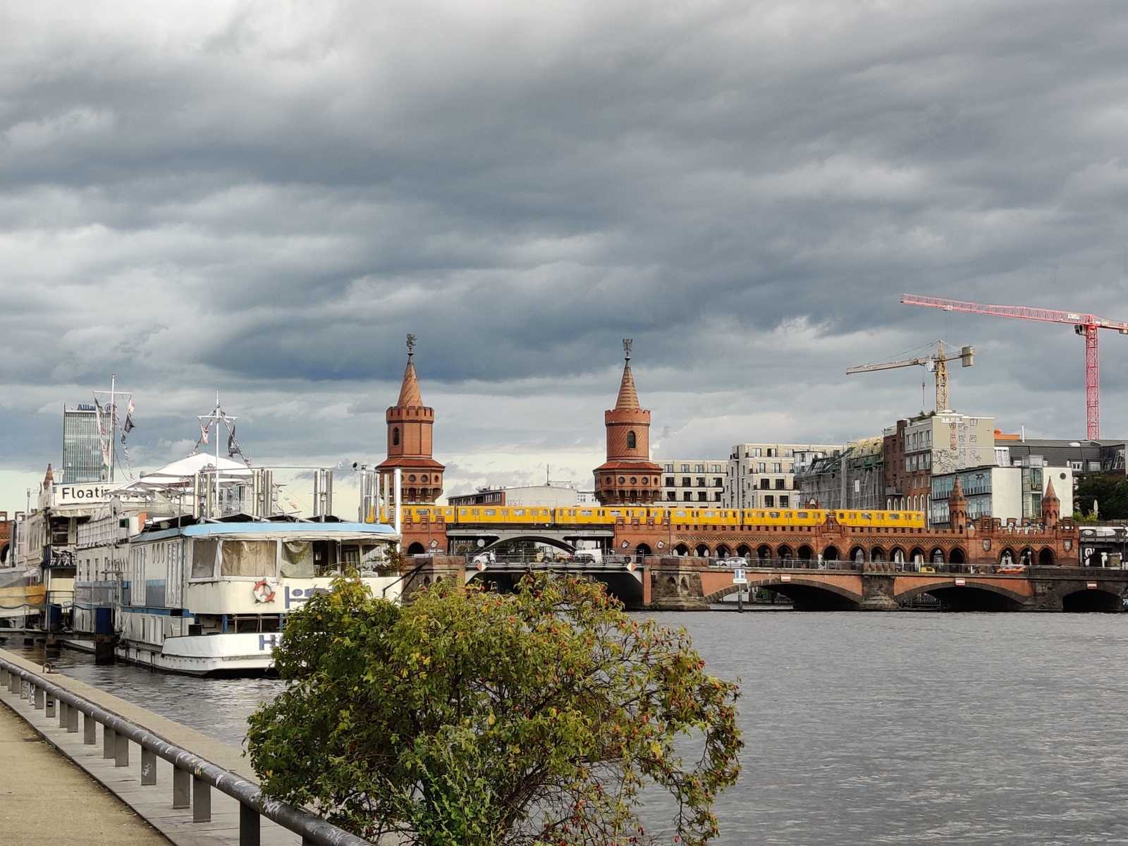 Blick auf die Oberbaumbrücke in Berlin, vom Spree-Ufer aus. Ein weißes Boot, welches eine Touristenunterkunft ist, liegt am Ufer an. Die gelben U-Bahnwägen der BVG ziehen auf der Brücke durch das Bild. Der Himmel ist von grauen Wolken bedeckt.
