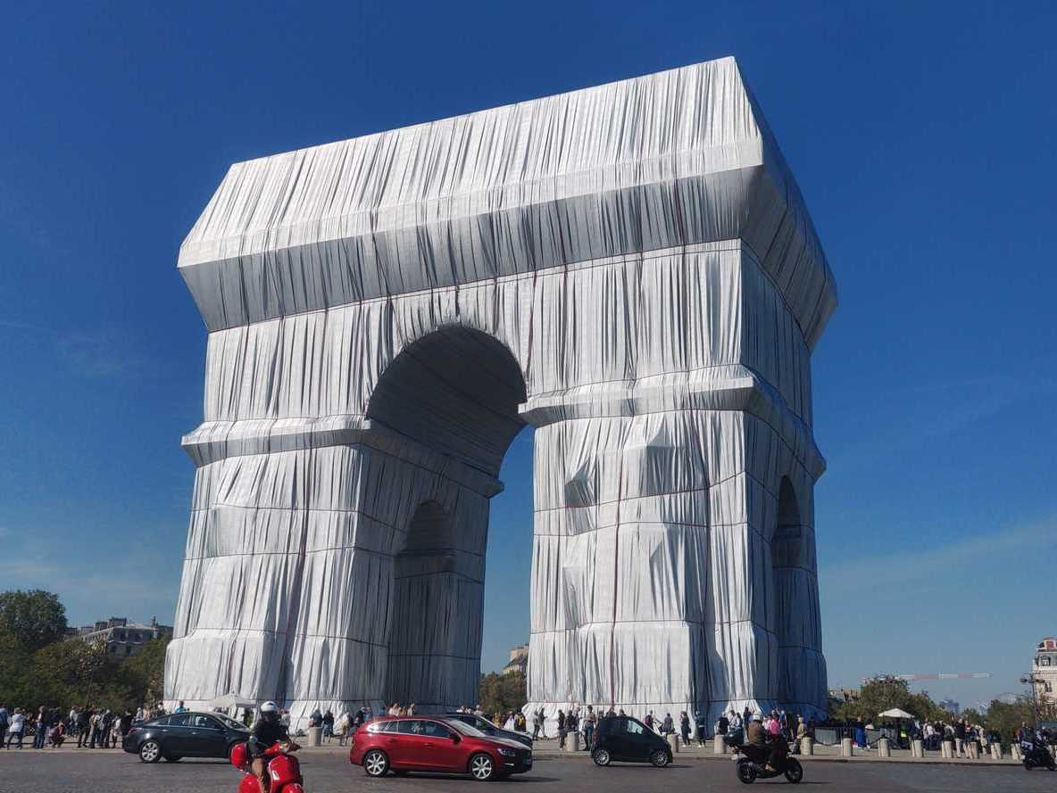 Paris, the shrouded Arc de Triomphe. Blue sky. Lots of car traffic. Wrapping art by Christo and Jeanne-Claude in a posthumous project. The fabric to wrap seems silver and leaves wrinkles.